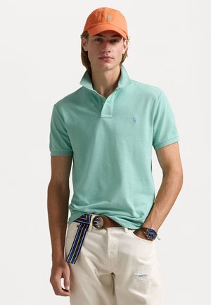 Muntgroen polo shirt met een kraag en een klein logo, gecombineerd met witte broek en een meerkleurige riem. Een oranje pet maakt de look compleet.