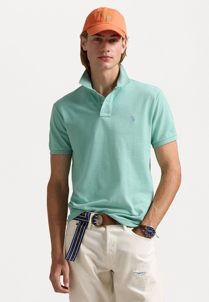 Muntgroen polo shirt met een kraag en een klein logo, gecombineerd met witte broek en een meerkleurige riem. Een oranje pet maakt de look compleet.