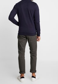 Mężczyzna w granatowej koszuli z długim rękawem, ciemnoszarych spodniach z podwiniętymi nogawkami i białych tenisówkach z czarnym detalem na pięcie, stojący na białym tle.