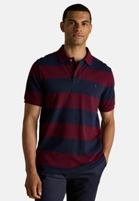 Polo krekls burgundijas un tumši zilo svītru dizainā, ar īsām piedurknēm, ar apkakli un mazu logo uz krūšu daļas. Izgatavots no kokvilnas materiāla ar gludu tekstūru.