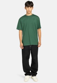 T-shirt in cotone verde con vestibilità rilassata, maniche corte e un piccolo dettaglio con logo. Abbinato a pantaloni in denim neri e sneakers bianche.