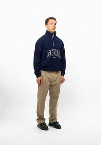 Navy-Fleece-Sweatshirt mit hohem Kragen und "GOOD"-Text. Beigefarbene Cordhosen und schwarze Schuhe. Glatte Textur und lässige Passform.