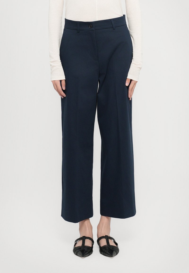 Marineblaue weit geschnittene Hose mit Knopfverschluss, die eine glatte Textur und eine maßgeschneiderte Passform aufweist. Begleitet von schwarzen Ballerinas mit Rippendetails.