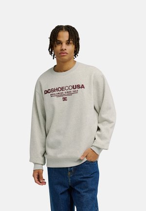 Junge Person mit kurzen Twists trägt ein hellgraues DC Shoes USA Sweatshirt und blaue Jeans, steht mit einer Hand in der Tasche und schaut nach vorne.