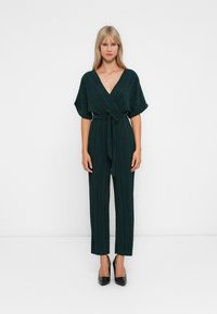 Kvinne som står mot hvit bakgrunn iført en mørkegrønn plissert jumpsuit med v-hals og belte, kombinert med svarte spisse hæler.