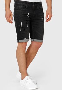 Pantaloni scurți negri din denim cu finisaj uzat, având rupturi și petice. Nasturi întorși și design clasic cu cinci buzunare. Asortați cu adidași albi.