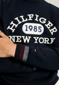 Granatowy sweter z haftem "Hilfiger 1985 New York" na piersi, ściągacze z czerwono-białymi oraz granatowymi paskami, małe logo na rękawie.