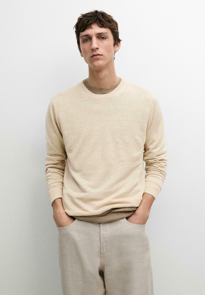 Jonge man met bruin haar draagt een beige trui in laagjes en lichtbeige broek, staand tegen een effen lichtgrijze achtergrond.