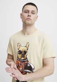 Lysegul bomulds T-shirt med et grafisk motiv af en hund i solbriller og en orange rumdragt, med sorte detaljer og en detaljeret illustration.