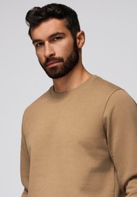 Homme aux cheveux foncés et à la barbe portant un sweat-shirt beige à col rond, tourné légèrement vers la gauche sur un fond clair uni.