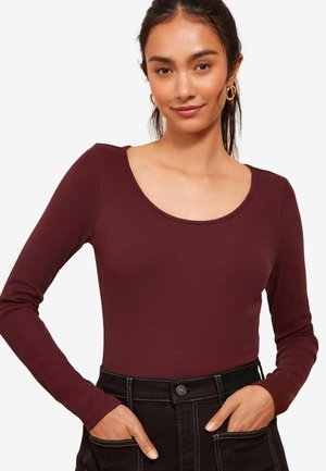 Bordeauxfarbene Langarmbluse mit Rundhalsausschnitt, gerippter Textur und tailliertem Schnitt, getragen zu hochgeschnittenen schwarzen Hosen mit kontrastierendem Nähten.