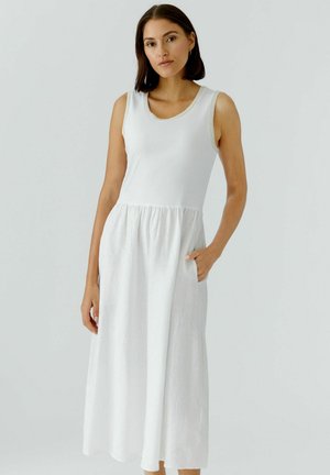 Junge Frau mit dunklen Haaren, die ein ärmelloses weißes Midikleid trägt und mit einer Hand in der Tasche vor einem schlichten hellen Hintergrund steht.