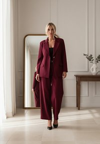 Bordowy garnitur z dopasowaną marynarką na jeden guzik i szerokimi spodniami, połączony z dopasowaną czarną bluzką i czarnymi czółenkami na szpic.