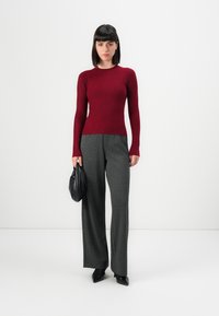 Femme portant un pull en rib de bourgogne ajusté, un pantalon large gris, des chaussures noires à bout pointu, tenant un sac à main en cuir noir, se tenant sur un fond blanc.