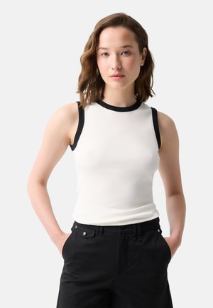 Vrouw met schouderlang bruin haar die een mouwloos wit topje met zwarte bies en zwarte broek draagt, staand met handen in de zakken.