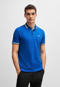 Sininen polo-paita, jossa on tasainen väri ja raidoitettu kaulus sekä hihansuut. Logo sijaitsee vasemmassa rinnassa. Materiaali näyttää sileältä.