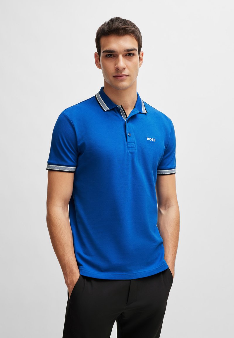 Sininen polo-paita, jossa on tasainen väri ja raidoitettu kaulus sekä hihansuut. Logo sijaitsee vasemmassa rinnassa. Materiaali näyttää sileältä.