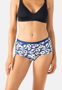Femme portant un bralette noir et un shorty en dentelle à motif floral bleu, se tenant devant un fond clair uni.