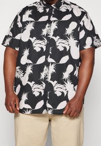 Korte mouwen shirt met een zwarte achtergrond en grote witte en lichtroze ananas- en bladdessins, met knoopsluiting aan de voorkant en een gladde stofstructuur.