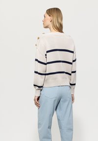 Beige Pullover mit marinefarbenen Streifen, gerippten Bündchen und goldenen Knöpfen an der Schulter, kombiniert mit hellblauen, locker sitzenden Hosen.