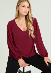 Bordowa bluzka z głębokim dekoltem w kształcie litery V, długimi rękawami i elastycznymi mankietami. Gładki materiał luźno opada na czarne spodnie.