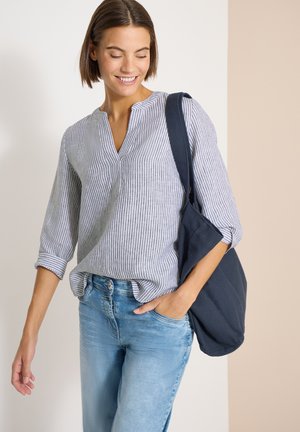 Joven mujer sonriendo, vistiendo una blusa gris a rayas y vaqueros azul claro, llevando un gran bolso de hombro azul oscuro.
