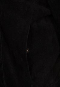 Binnenkant van een zwart suède jack, met een gladde textuur en een zichtbaar knoopdetail. De stof lijkt zacht met een matte afwerking.