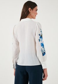Blouse blanche à manches bouffantes, avec une broderie florale bleue sur la manche gauche et un col classique. Tissu doux et léger.