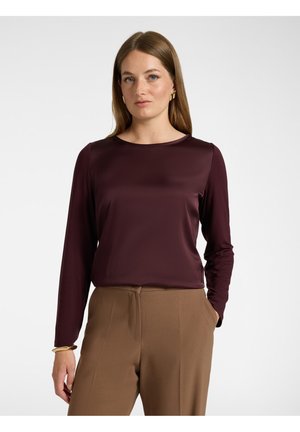 Frau mit glatten braunen Haaren, die ein langärmliges dunkelviolettes Oberteil und braune Hose trägt, steht mit einer Hand in der Tasche, neutraler Hintergrund.