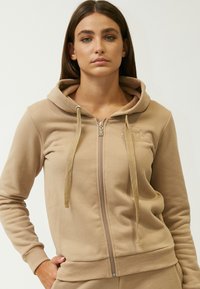 Felpa con zip di colore beige, dotata di lacci e tasca a marsupio. Tessuto morbido, polsini e vita a coste. Presenta un piccolo logo sul petto.