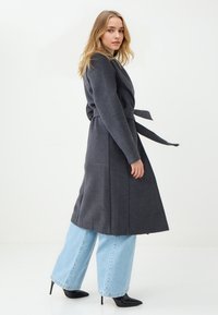 Lang grijze jas met een wikkelontwerp, ceintuur in de taille en zijzakken, gecombineerd met lichtblauwe jeans met wijde broekspijpen en zwarte puntige hakken.