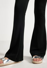 Benen in zwarte uitlopende broeken en zilveren gespsandalen met beige dikke zolen, staande op een lichte vloer tegen een effen achtergrond.