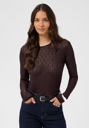 Jeune femme aux longs cheveux ondulés porte un haut sombre transparent à manches longues rentré dans un jean foncé avec une ceinture noire, les mains dans les poches avant.