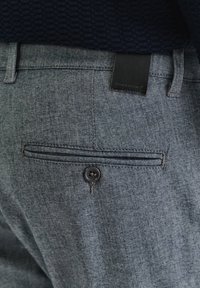 Ciemnoniebieskie materiałowe spodnie z tylną kieszenią, zapięciem na guzik i strukturalnym pasem w średnim splocie denimu. Widoczna metka akcentowa.