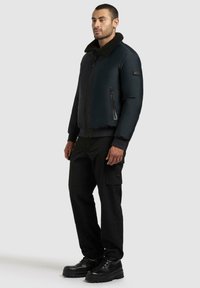 Sort bomberjakke med teksturert krage, glidelåsfeste foran og sidelommer, kombinert med svarte cargobukser og kraftige svarte støvler.