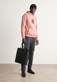 Sudadera con capucha rosa con el logo "HUGO", bolso negro de mano con dos asas, jeans grises y zapatos negros, llevados por una persona.