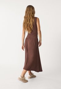 Ruskea hihaton mekko, jossa on istuva päälliosa ja levenevä hameosa, ja vyötäröllä on rypytetty muotoilu. Yhdistetty vaaleanruskeisiin tekstuuripintoihin lenkkareihin.