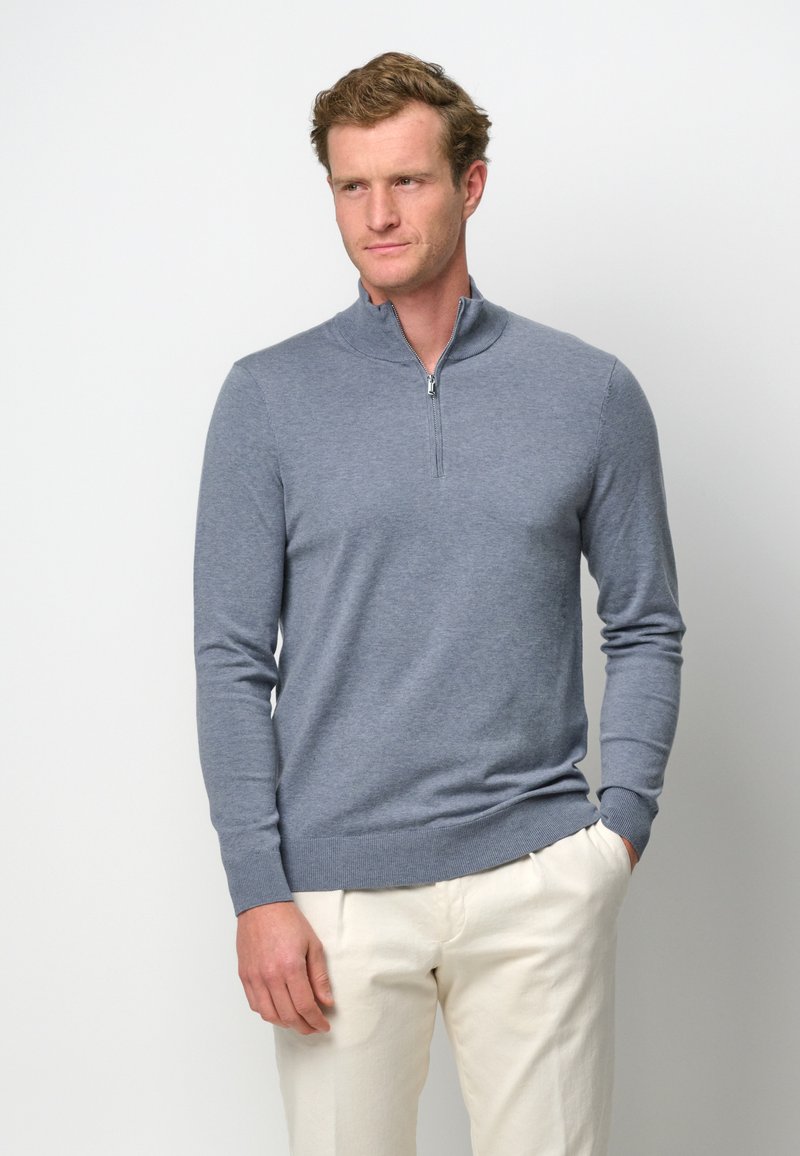 Grauer Zip-Pullover aus weichem Strickmaterial mit langen Ärmeln sowie gerippten Bündchen und Saum, kombiniert mit hellen Hosen.