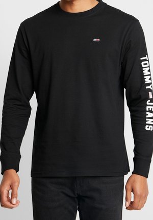 Černé tričko s dlouhým rukávem vyrobené z bavlny, s malým logem na hrudi a nápisem "TOMMY JEANS" vytištěným na levém rukávu bílou barvou.