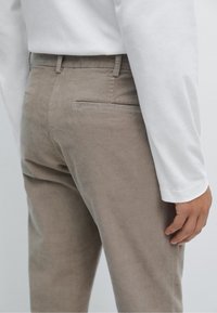 Pantaloni di velluto a coste beige con una texture liscia, dotati di una tasca posteriore e di una vita. Indossati con una camicia bianca a maniche lunghe.