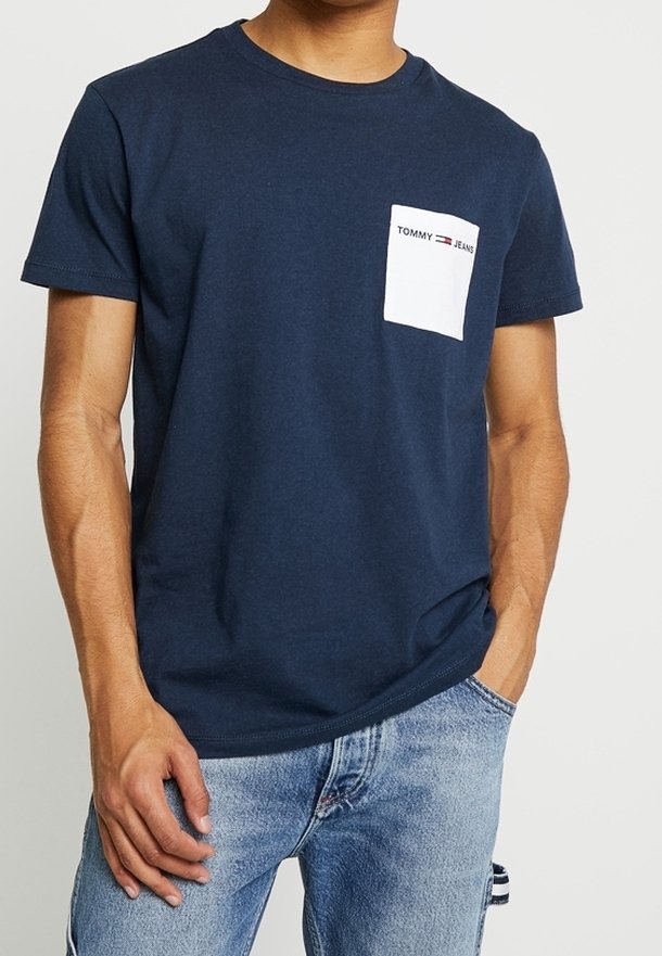 T-paita tummansinistä puuvillaa, jossa on valkoinen rintatasku, jossa on "TOMMY JEANS" -logo, pyöreä kaula-aukko ja lyhyet hihat.