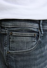Tumši mazgāti džinsi ar šauru piegriezumu. Iespējas ietver aizmugurējo kabatu ar šuvju detaļām un metāla kniedēm. Audums ir gluds ar nelielu nodilumu.