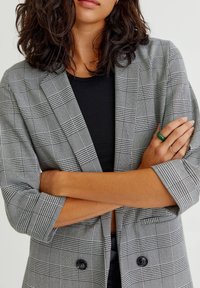 Blazer a quadri grigi con una vestibilità rilassata, maniche arrotolate e due bottoni neri. Indossato sopra una camicia nera che mostra un motivo a scacchi.