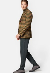 Brązowa i zielona marynarka w kratę, oliwkowy golf, grafitowe spodnie i szare buty. Marynarka ma klapy oraz boczne kieszenie.