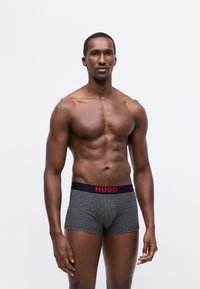 Boxeri negri cu un model repetitiv "HUGO" în gri, având o talie solidă neagră cu un logo roșu îndrăzneț "HUGO" în față.