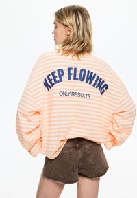 Prevelika majica v breskvaste črte z modrim napisom "KEEP FLOWING ONLY RESULTS" na hrbtni strani; kombinirana z rjavimi denim kratkimi hlačami. Mehka tkanina z udobnim krojem.
