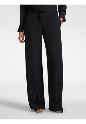 Sorte bukser med vide ben og broderede svirler nær kanten, båret med guldglitrende sko og guldarmbånd, den ene hånd i lommen.