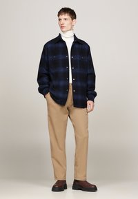 Navy plaid shirt with two chest pockets, paired with beige trousers and brown slip-on boots. Smooth fabric with a relaxed fit.