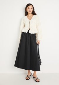 Biela textúrovaná blúzka s hlbokým výstrihom do V a dlhými rukávmi, v kombinácii s čiernou maxi sukňou a čiernymi sandálmi na podpätku. Čierna taška so popruhmi.