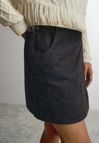 Denim miniskirt i mørkebrun med en struktureret overflade, der har en sidelomme, kontrasterende syninger og en knapdetalje i taljen.